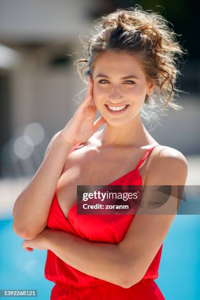 beautiful young fashion model wearing red sleeveless dress posing by the pool and smiling at camera - décolleté stock pictures, royalty-free photos & images