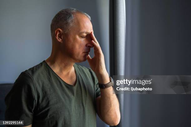 orried mature man standing at home, close to a window, with hand in the head - pain imagens e fotografias de stock