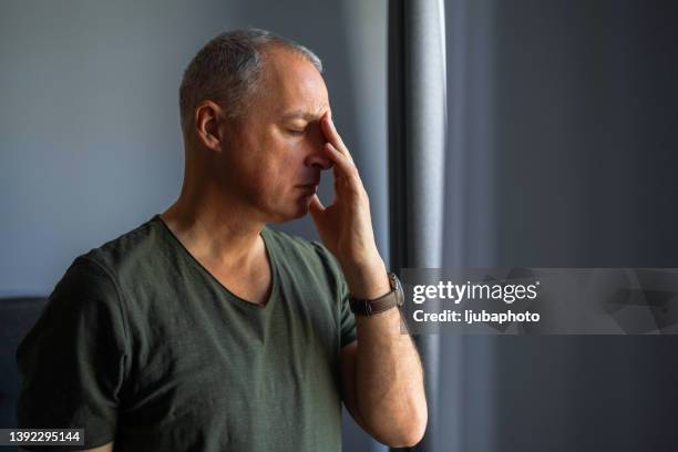 orried mature man standing at home, close to a window, with hand in the head - sinusitis 個照片及圖片檔