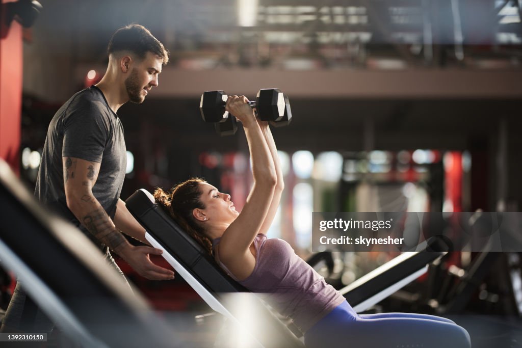 Junger Mann, der seiner Freundin beim Sporttraining in einem Fitnessstudio hilft.