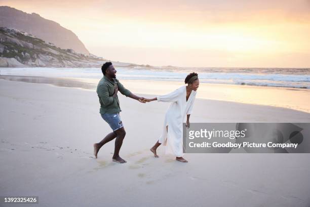 pareja joven riendo mientras corren juntos por una playa de arena al atardecer - luna de miel fotografías e imágenes de stock