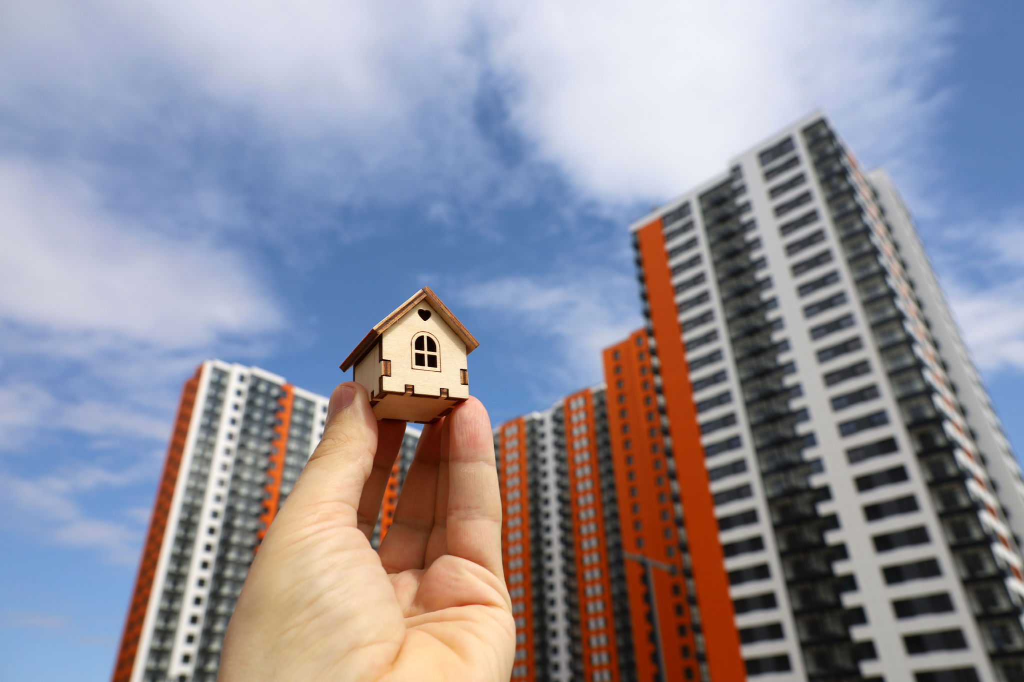 Wooden house model in male hand on background of new buildings and blue sky Wooden house model in male hand on background of new buildings and blue sky