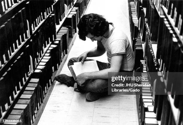 Inside the Milton S Eisenhower Library at Johns Hopkins University, a male student wearing a tee shirt and jeans sits on the floor between two stacks...