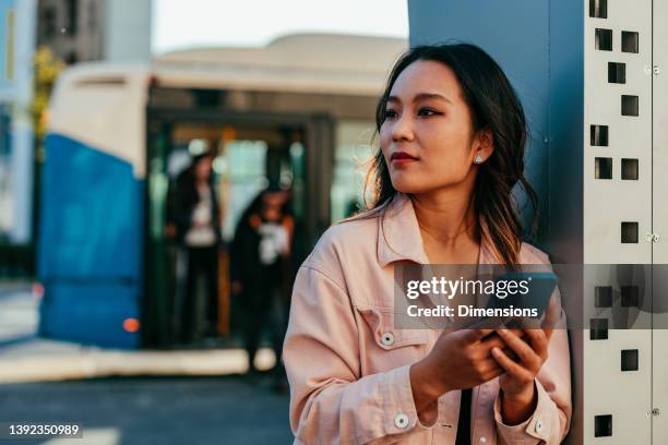 young woman holding cellphone while waiting for bus - bus station stock pictures, royalty-free photos & images