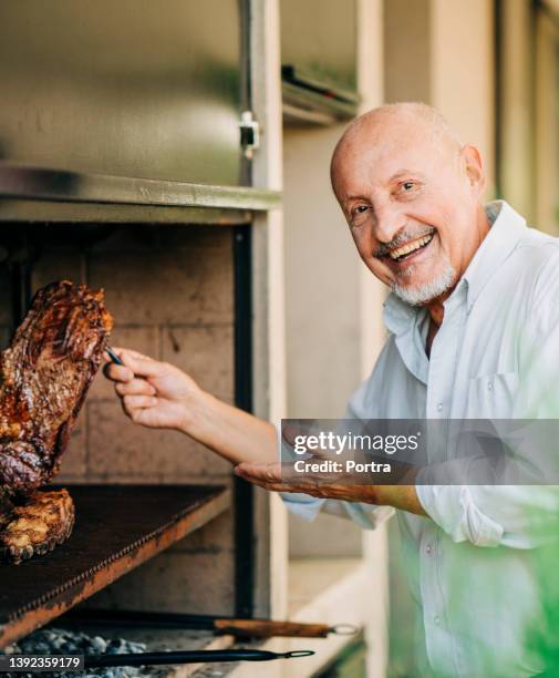 lächelnder älterer mann, der fleisch auf grill zubereitet - asado stock-fotos und bilder