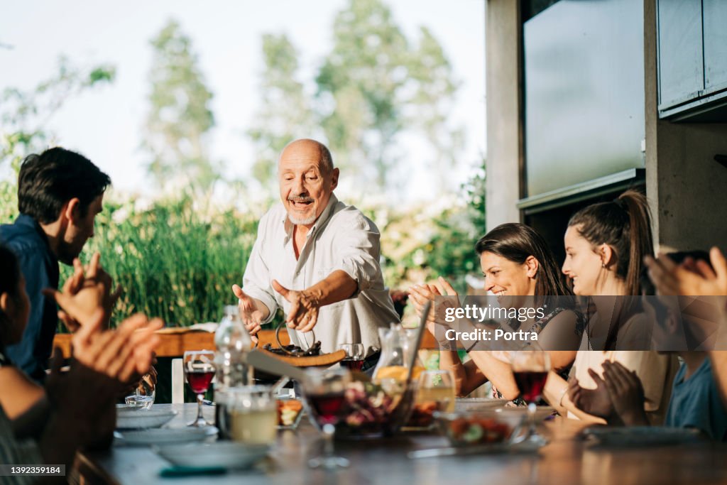 Cheerful senior man serving barbecue meat to family sitting in backyard dining table