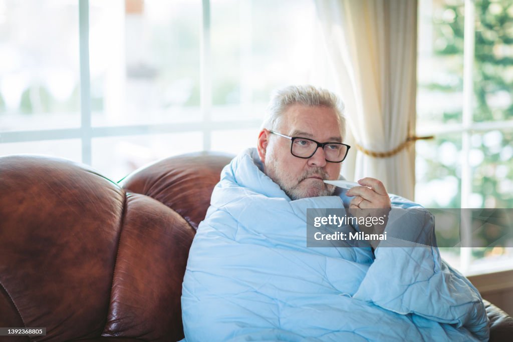 Cold exhausted senior man with flu wrapped in a warm blanket. He is holding a thermometer.