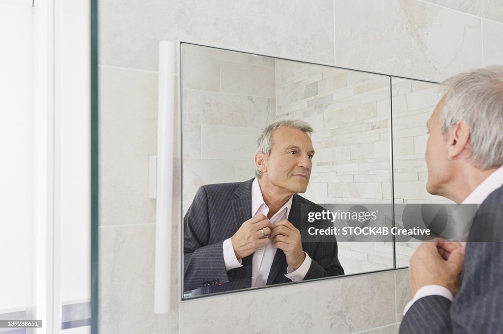 Senior man looking in bathroom mirror buttoning his tie