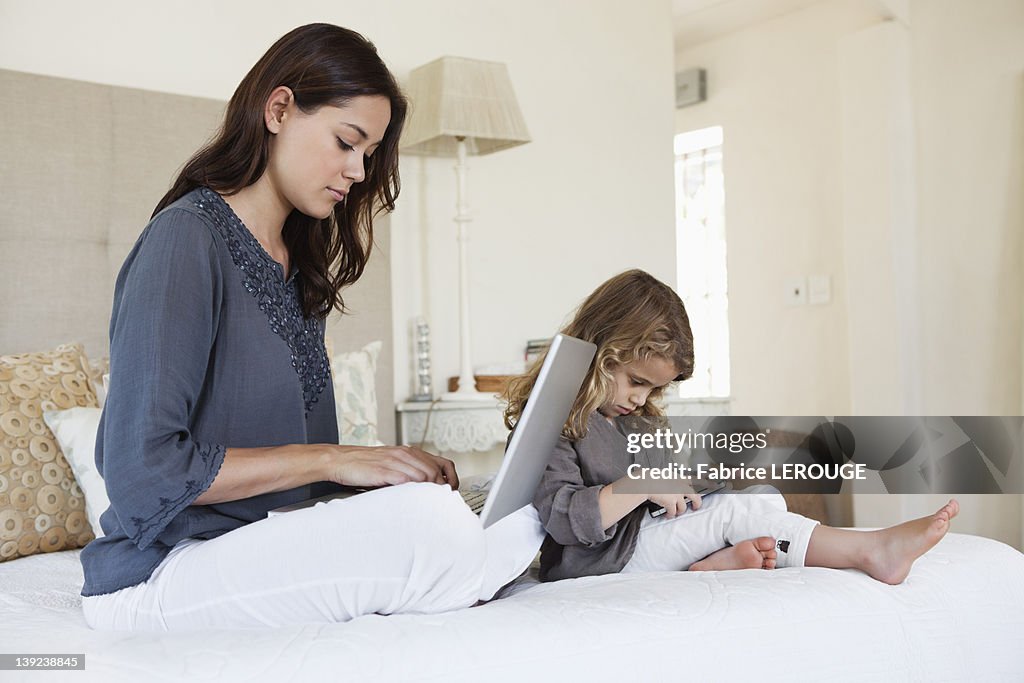 Woman working on a laptop with her daughter playing with a mobile phone beside her