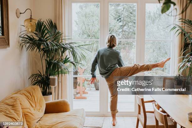 a flexible man stretches / dances in front of large patio doors in a stylish home environment - standing on one leg stock-fotos und bilder