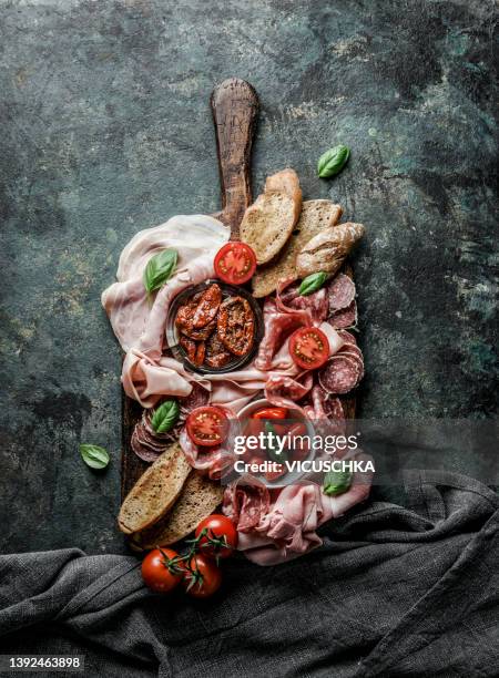 italian meat plate with various ham, salami, ciabatta bread, tomatoes and basil leaves on wooden cutting board on rustic dark kitchen table background. - antipasto stock-fotos und bilder