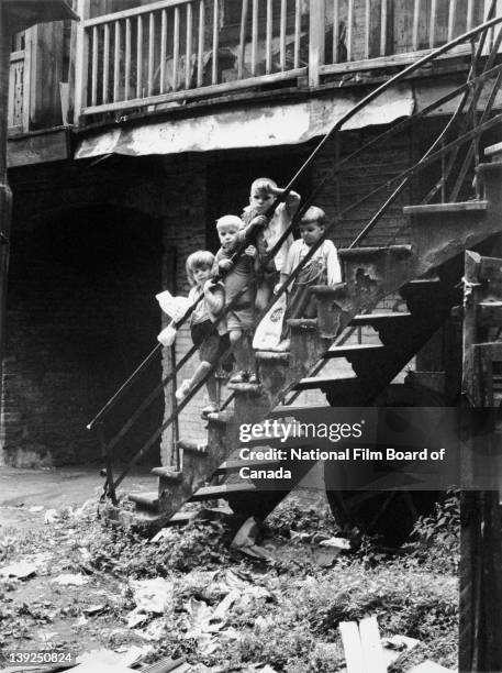 Children stand on the backyard staircase of a rundown tenement building in the slum neighborhood of a city, Canada, 1968. Photo taken during the...