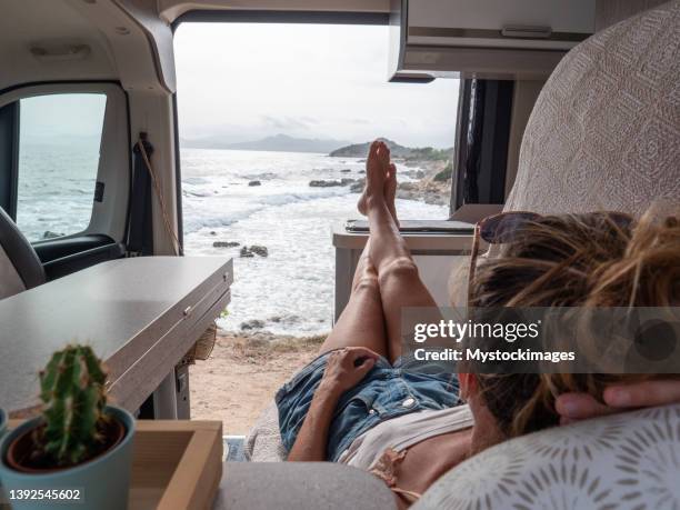 woman in her camper van looking at ocean, she lies on the bench, side door open - van life stock pictures, royalty-free photos & images