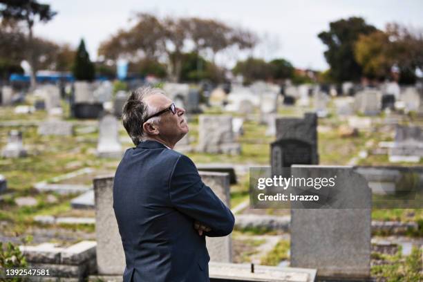 a middle-aged man stands in front of graves in a cemetery looking up. - funeral planning stock pictures, royalty-free photos & images