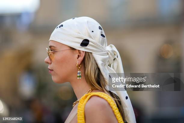 Almuneda Lapique wears yellow sunglasses, gold and blue / green pearls pendant earrings, a white with black print pattern silk scarf as a headband, a...