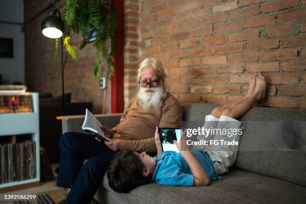 grandfather reading and book and grandson using digital tablet at home - generation gap stock pictures, royalty-free photos & images