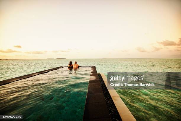 extreme wide shot of mature couple watching sunrise from infinity pool - resort swimming pool stockfoto's en -beelden