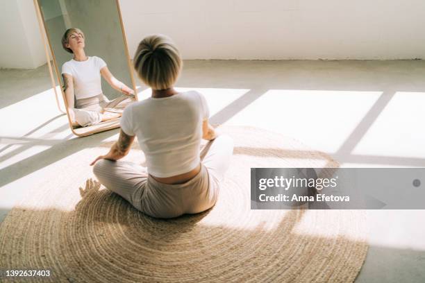 beautiful authentic woman with tattoos and short blond hair is meditating sitting in lotus position on wicker carpet in front of a mirror. she is wearing a light-colored casual clothing. concept of relaxation exercises - schneidersitz stock-fotos und bilder