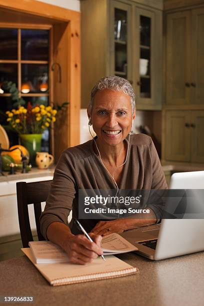mulher madura com computador portátil e vestindo retrato de ouvido telefones, - 50s woman writing at table imagens e fotografias de stock
