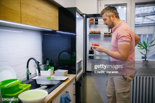 homme souriant et fermant la porte du réfrigérateur, tenant une boîte de tomates cerises - réfrigérateur fermé photos et images de collection