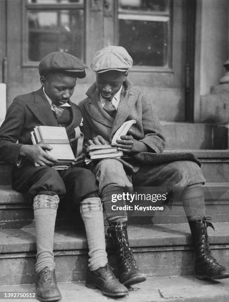 Two boys wearing knickerbockers and baker boys caps sit on a flight of steps, one boy holding a bundle of books while the other holds open a book...