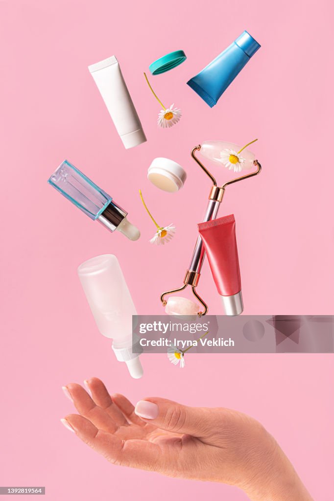 Woman hand holds flying skin care products on pastel pink color background.