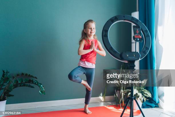little girl standing on one leg with joined hands namaste standing in tree pose yoga while phone shooting video of training - yoga studio shot stock pictures, royalty-free photos & images