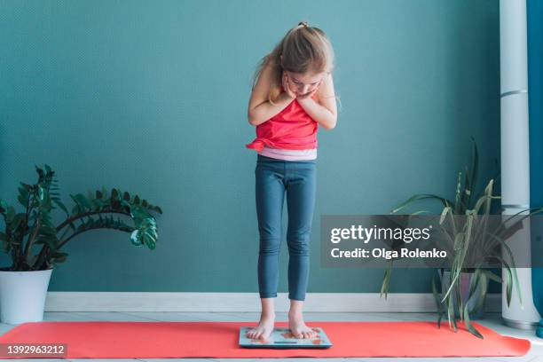 cute little barefoot girl standing on weight scale. holding head in hands. gain weight. - fat loss training stockfoto's en -beelden