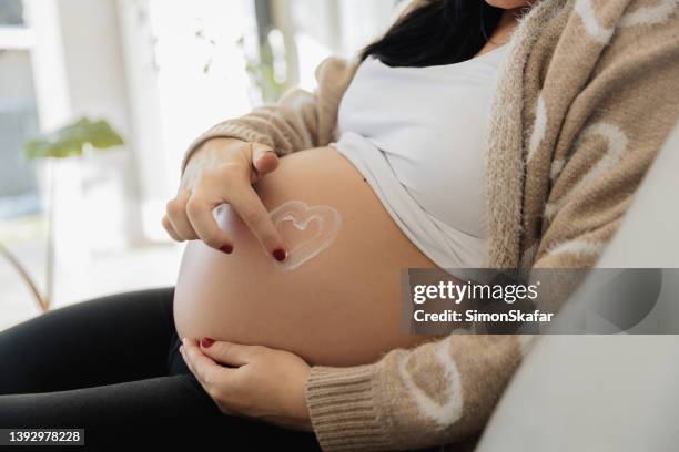 pregnant woman drawing heart with finger on her belly on sofa - pregnant belly stock pictures, royalty-free photos & images