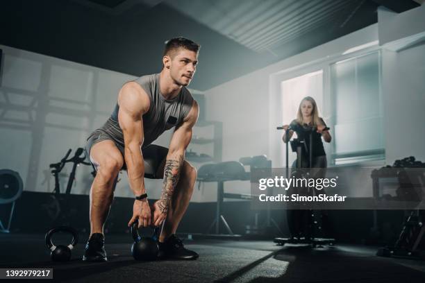 male athlete training with kettlebell while female cycling on exercise bike at health club - crossfit stock pictures, royalty-free photos & images