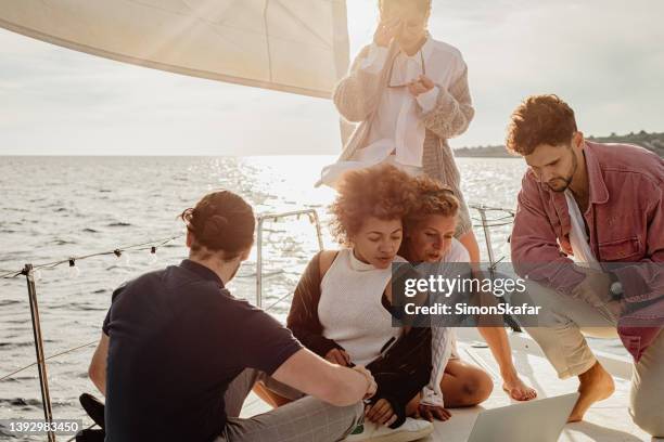 friends sitting close to each other on boat deck of a sailboat in the evening looking at a laptop, sunlight in background - sail boat deck stock pictures, royalty-free photos & images