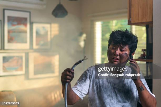 portrait of young man holding electrical cable after domestic accident with dirty burnt funny face expression calling desperate with mobile phone asking for help. electricity repairs and diy concept. - diy disaster stock pictures, royalty-free photos & images