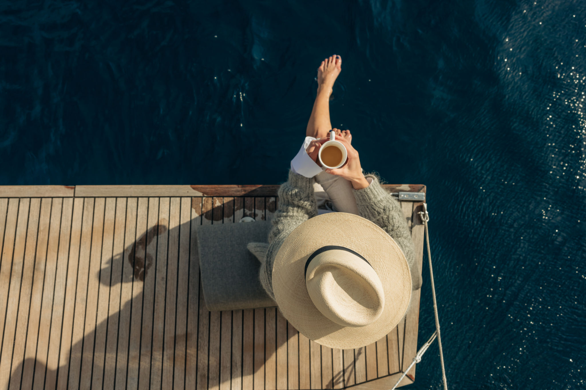 Women with hat holding a cup of coffee in the morning, sitting on back boat deck of a sailboat, bar feet legs in the water, view from high angle Women with hat holding a cup of coffee in the morning, sitting on back boat deck of a sailboat, bar feet legs in the water, view from high angle
