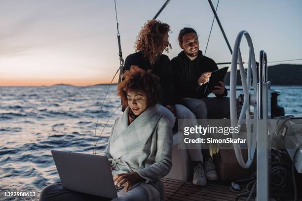 two women with curly hair and a man sitting on boat deck of a sailboat in the evening working on a laptop and looking at a tablet - sail boat deck stock pictures, royalty-free photos & images