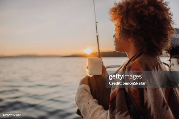 日の出時にボートデッキに座りながら海を眺める茶色の巻き毛の女性で、温かい飲み物を飲みながら - hot women on boats ストックフォトと画像