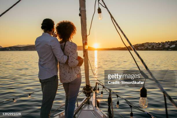man embracing woman, both standing on front boat deck of a sailboat in the evening, looking at the sunset above an island, rear view - sail boat deck stock pictures, royalty-free photos & images