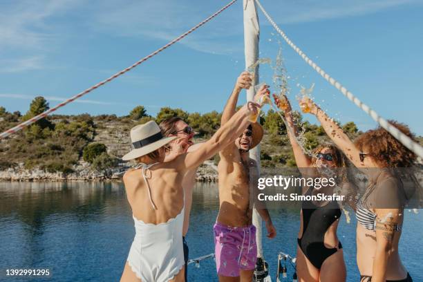 group of friends with swimwear smiling and laughing while toasting beer on boat deck of a sailboat, beer and liquid flies around, cheerful mood - beer splash stock pictures, royalty-free photos & images