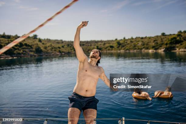homem com longos cabelos castanhos amarrados em uma trança e barba pulando no mar do convés de um veleiro enquanto tirava uma selfie com o telefone - mens long jump - fotografias e filmes do acervo