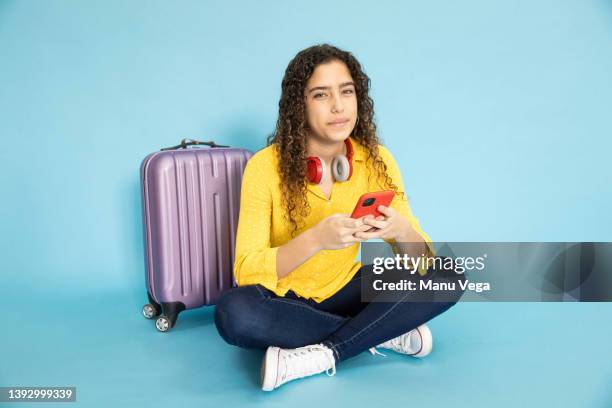 young traveler sitting on floor with her luggage holding her cell phone and looking at camera - young woman trolley stock-fotos und bilder
