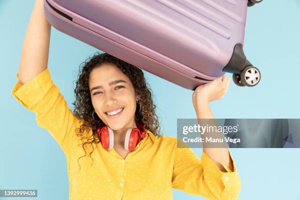 young woman lifting her wheeled suitcase while looking at camera - carry on bag imagens e fotografias de stock