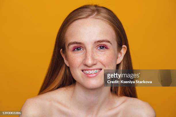 a young beautiful european woman with blond hair and freckles, multi-colored mascara and makeup on a yellow background looks at the camera and smiles, on her teeth there are skyce rhinestones - strass imagens e fotografias de stock