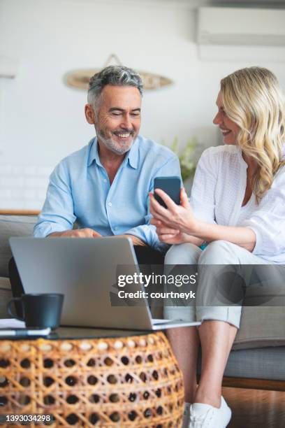 mature couple using a laptop computer and mobile phone on the sofa - person looking at phone while smiling australia stock pictures, royalty-free photos & images