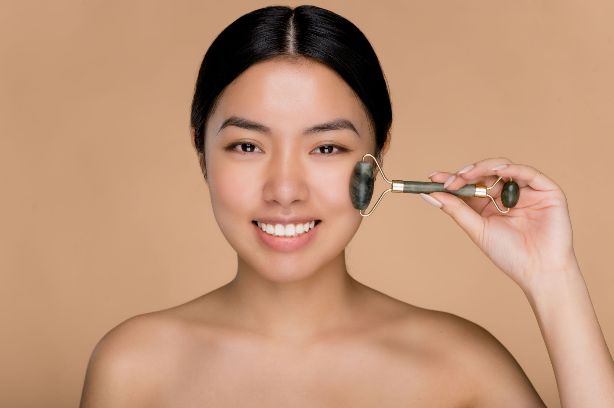 Close up photo of charming beautiful young Asian woman with naked shoulders, using derma roller for massage her facial skin. Photo of a smiling woman on isolated beige background Close up photo of charming beautiful young Asian woman with naked shoulders, using derma roller for massage her facial skin. Photo of a smiling woman on isolated beige background