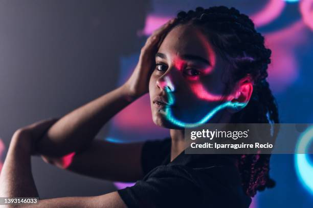 a young woman with braids in a nightclub. room lit in retro/retrowave style with neon lights and pink and blue projections. making fashion poses with your hands - disco costume stockfoto's en -beelden