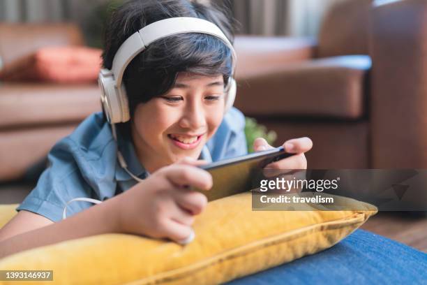 chilling weekend holiday with esport game online,asian child boy wear headphone hand playing smartphone game online with exited and fun laydown on floor at living room at home, son is playing alone with mobile equipment. - home alone movie stockfoto's en -beelden