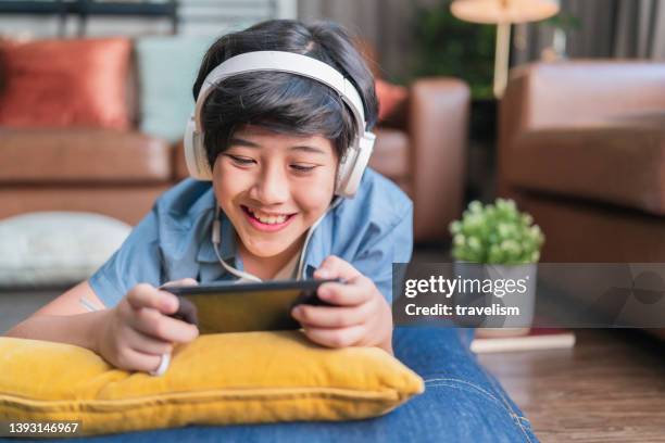 chilling weekend holiday with esport game online,asian child boy wear headphone hand playing smartphone game online with exited and fun laydown on floor at living room at home, son is playing alone with mobile equipment. - home alone movie stockfoto's en -beelden