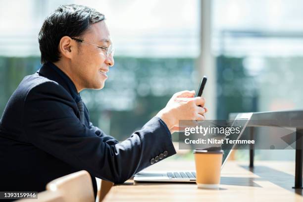 smart technology for business entrepreneur working anywhere. side view of  a japanese business entrepreur in a hotel lobby while reading information on mobile phone or checking business calendar to making appointment with his team. - ノートパソコン スマートフォン ストックフォトと�画像