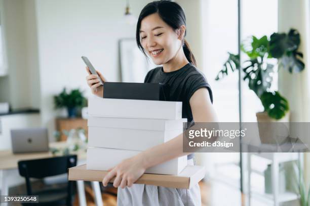 happy and satisfied young asian woman with a stack of cardboard boxes delivery package in her arms using smartphone to rate the products and service, sending positive impression. online shopping. trustworthy delivery service concept - debit cards credit cards accepted stock pictures, royalty-free photos & images