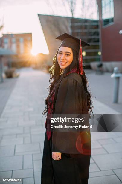 portrait of a cheerful young hispanic woman wearing a cap and gown looking over her shoulder with a maroon sash in the spring before graduation - maroon graduation stock pictures, royalty-free photos & images