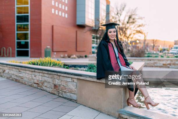 portrait of a cheerful young hispanic woman wearing a cap and gown with a maroon sash, sitting by a fountain - maroon graduation stock pictures, royalty-free photos & images
